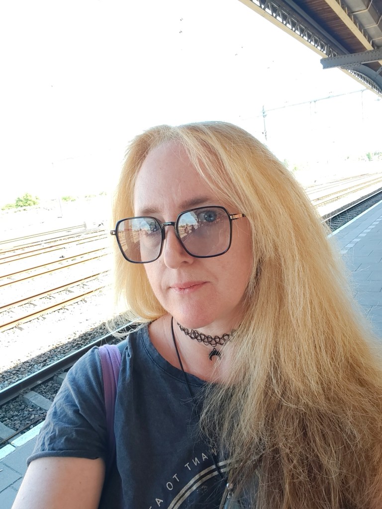 A person with long, blonde hair wearing large glasses and a choker necklace, standing on a train platform with railway tracks in the background on a sunny day.