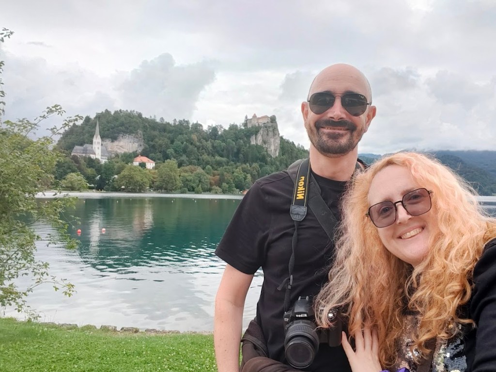 Couple taking selfie by calm lake with historic castle in background, lush green landscape, and dramatic cloudy sky.