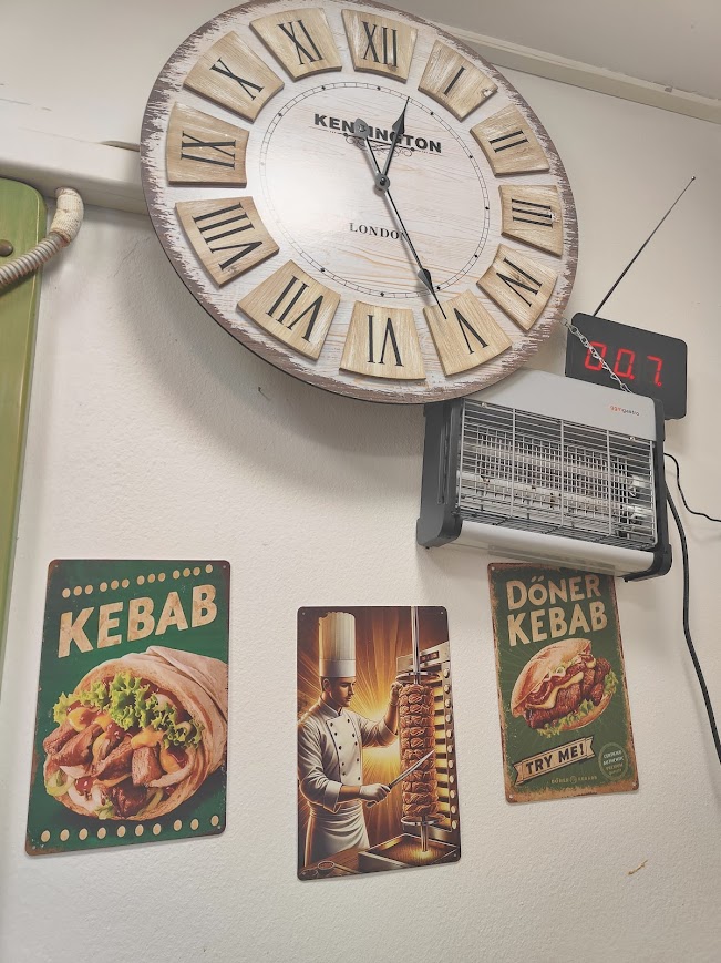 Restaurant wall with large wooden clock, vintage kebab signs including lettuce‑wrapped kebab, chef slicing meat from rotisserie, and Döner Kebab dish illustration, with wall heater.