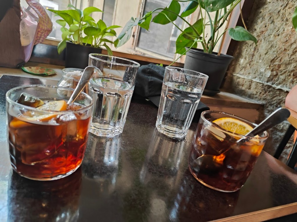 Close‑up of two iced cocktails with lemon slices on dark table, served with water glasses and green plant in background.
