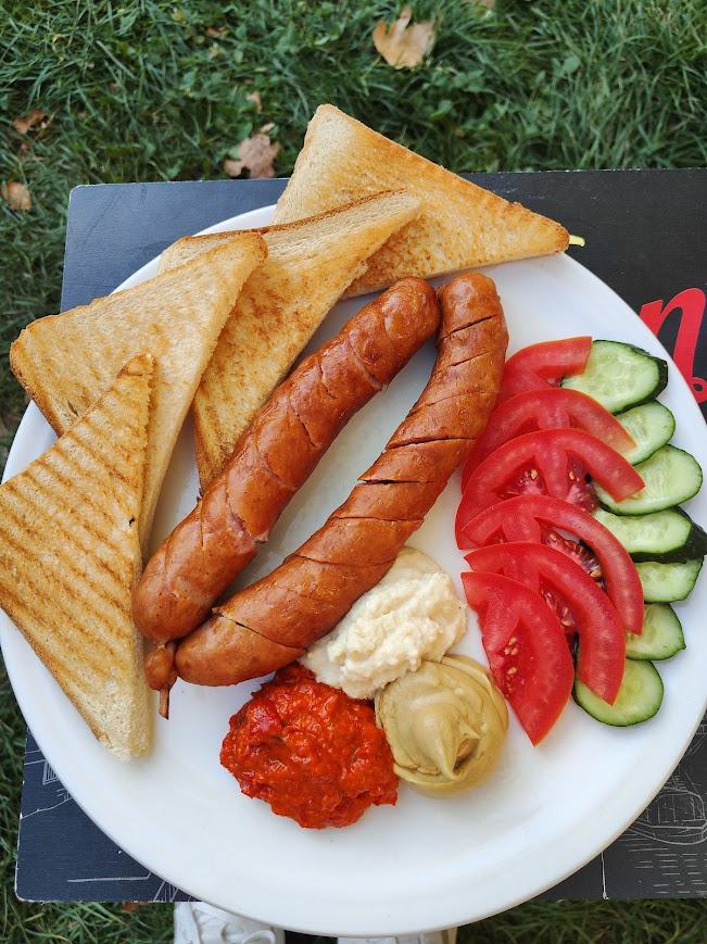 Plate of grilled sausages with toasted bread, fresh tomato and cucumber slices, and assorted dipping sauces.