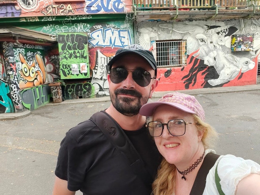 Couple taking selfie in front of colorful graffiti‑covered wall featuring vibrant urban street art.