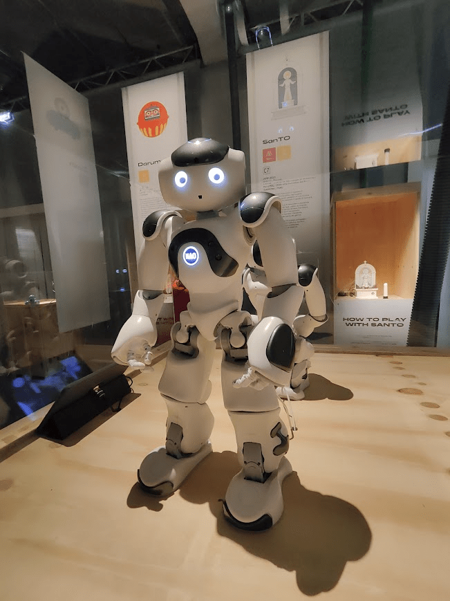 A white humanoid robot with glowing blue eyes stands in a display area, surrounded by informational panels.