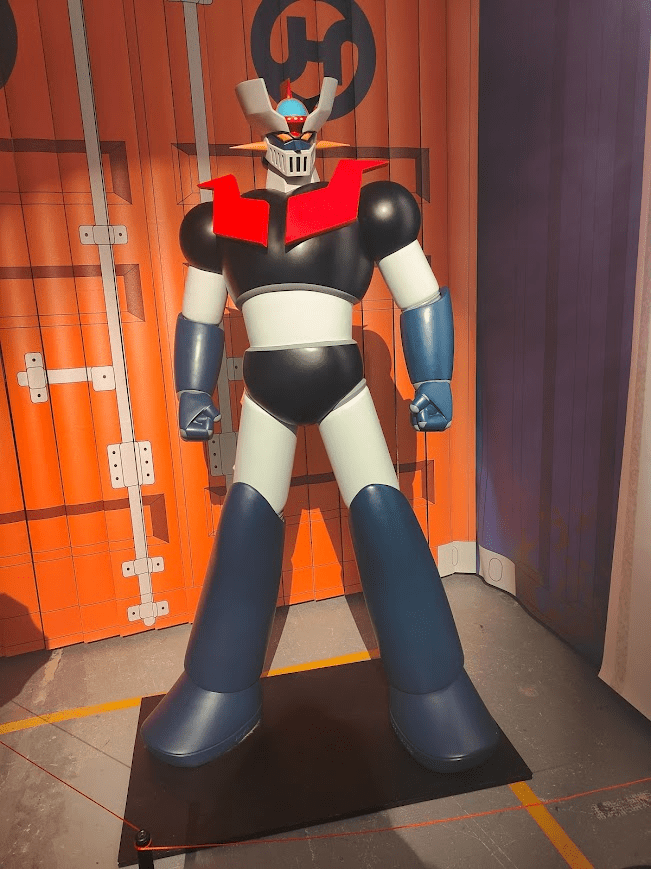 A large, stylized Mazinger Z figure with a black and white body, red shoulders, and a helmet with antennae, standing on a black base against a backdrop of orange and purple walls.