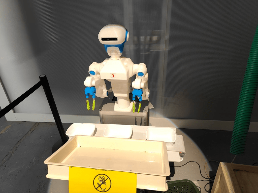 A robotic arm with blue and white colors standing next to white trays on a table, with a caution sign indicating safety protocols.